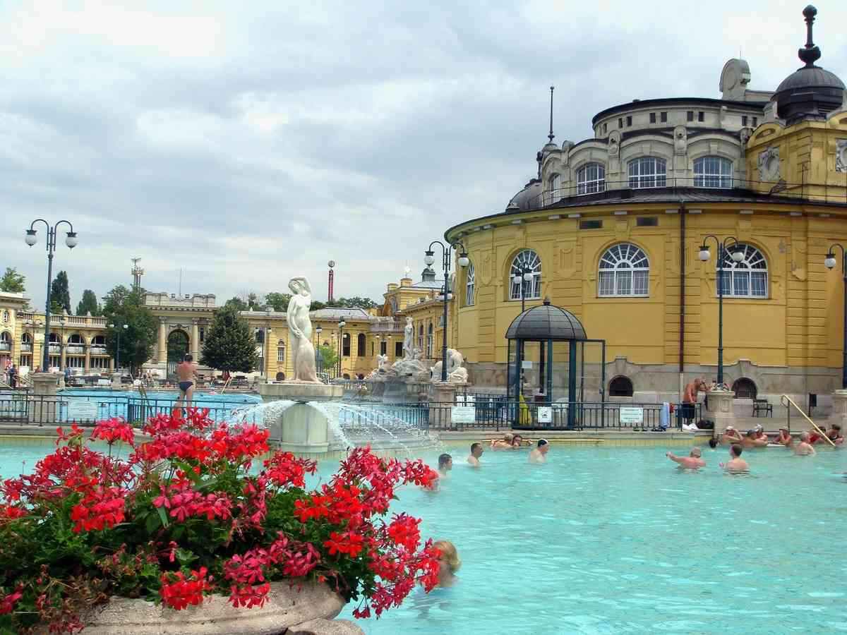 Piscine thermale turquoise entourée d'architecture jaune, baigneurs, fleurs rouges, bâtiments historiques